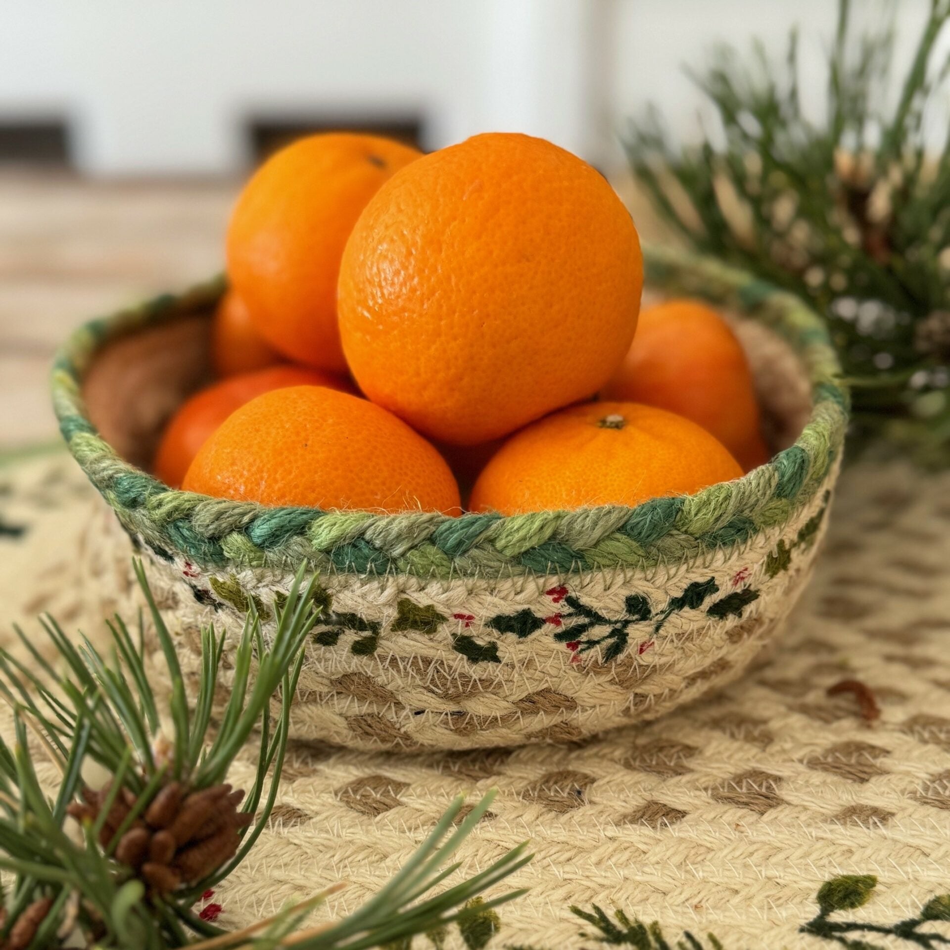 A jute bowl with a green and cream embroidered design holds several bright orange mandarins.
