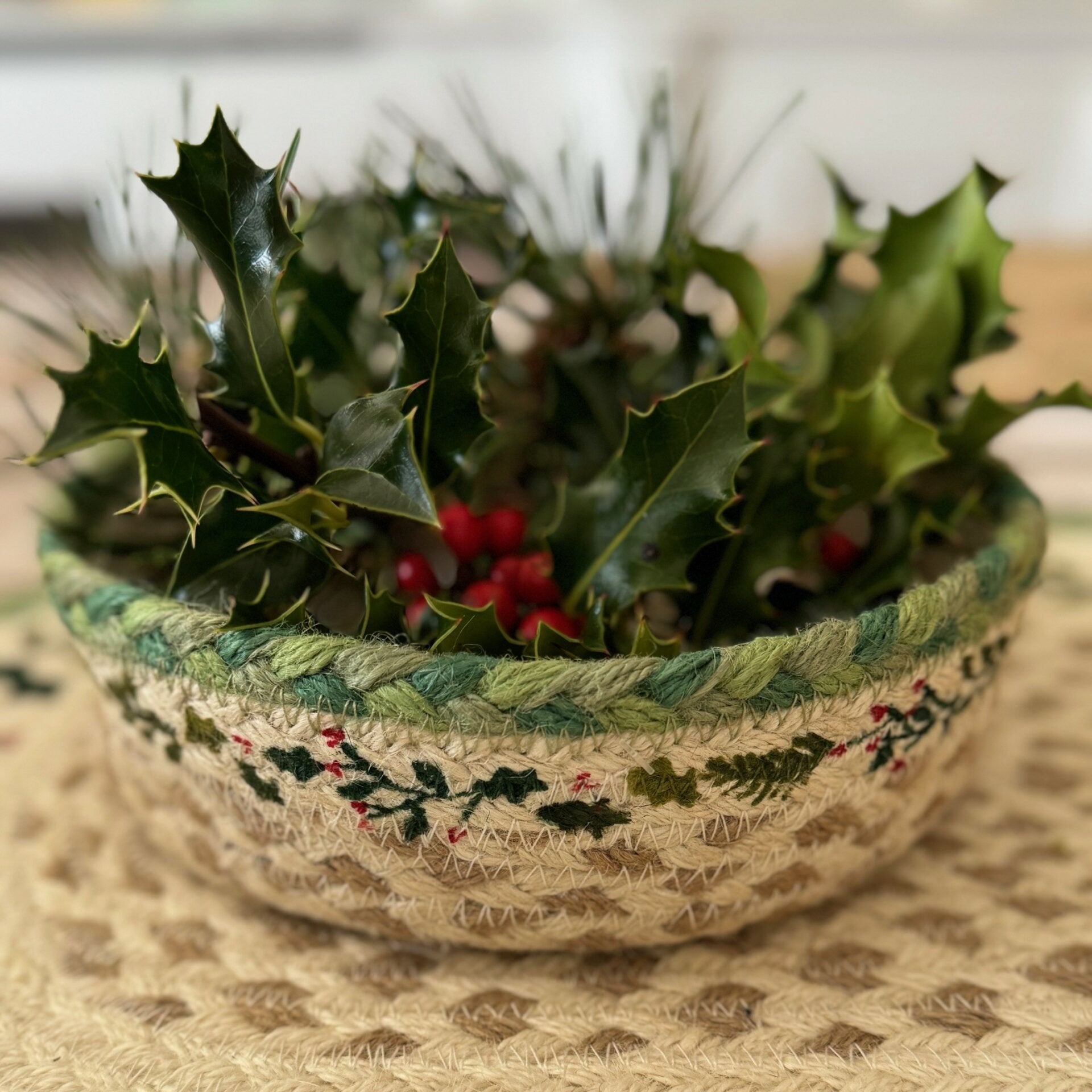 A beige jute bowl with green and red embroidered holly sprigs, filled with fresh holly sprigs and red berries.