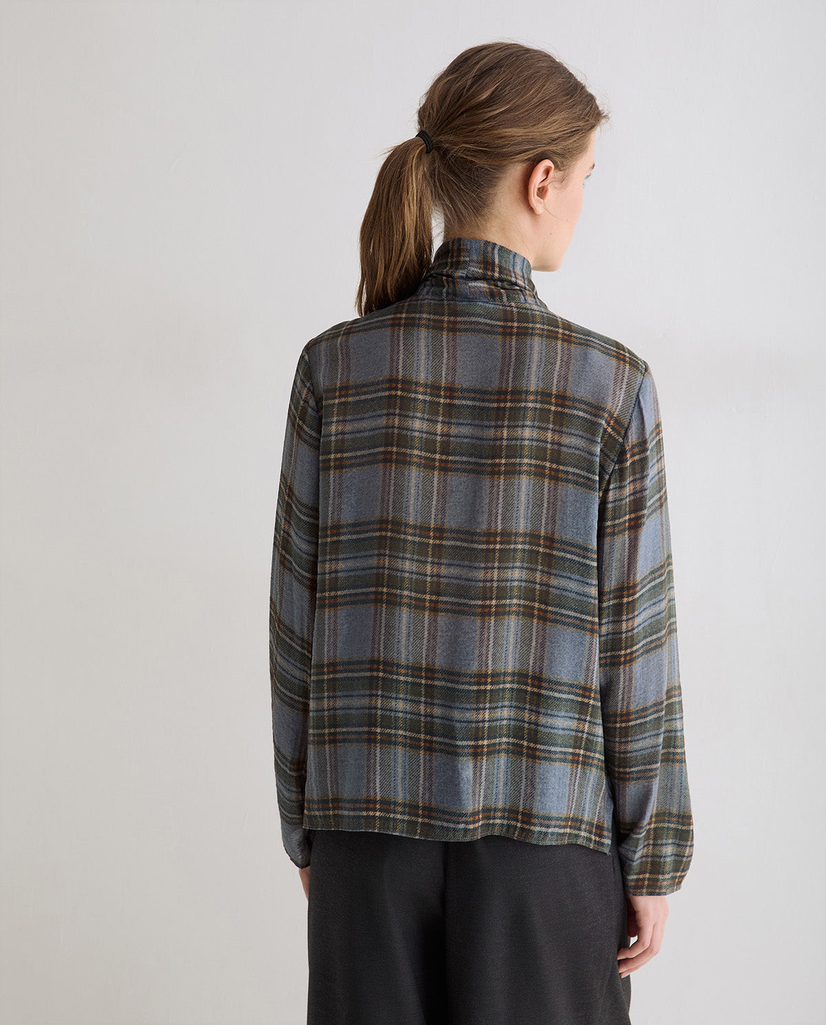 A woman viewed from the back wearing a long-sleeve plaid shirt in muted blue-gray, brown, and beige tones, paired with dark pants, against a plain light gray studio background.
