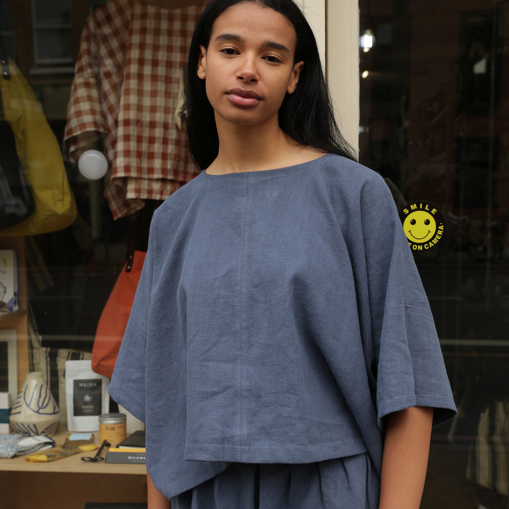 Dusty blue linen top with short sleeves and a boxy fit, featuring a small "Smile on Camera" graphic.