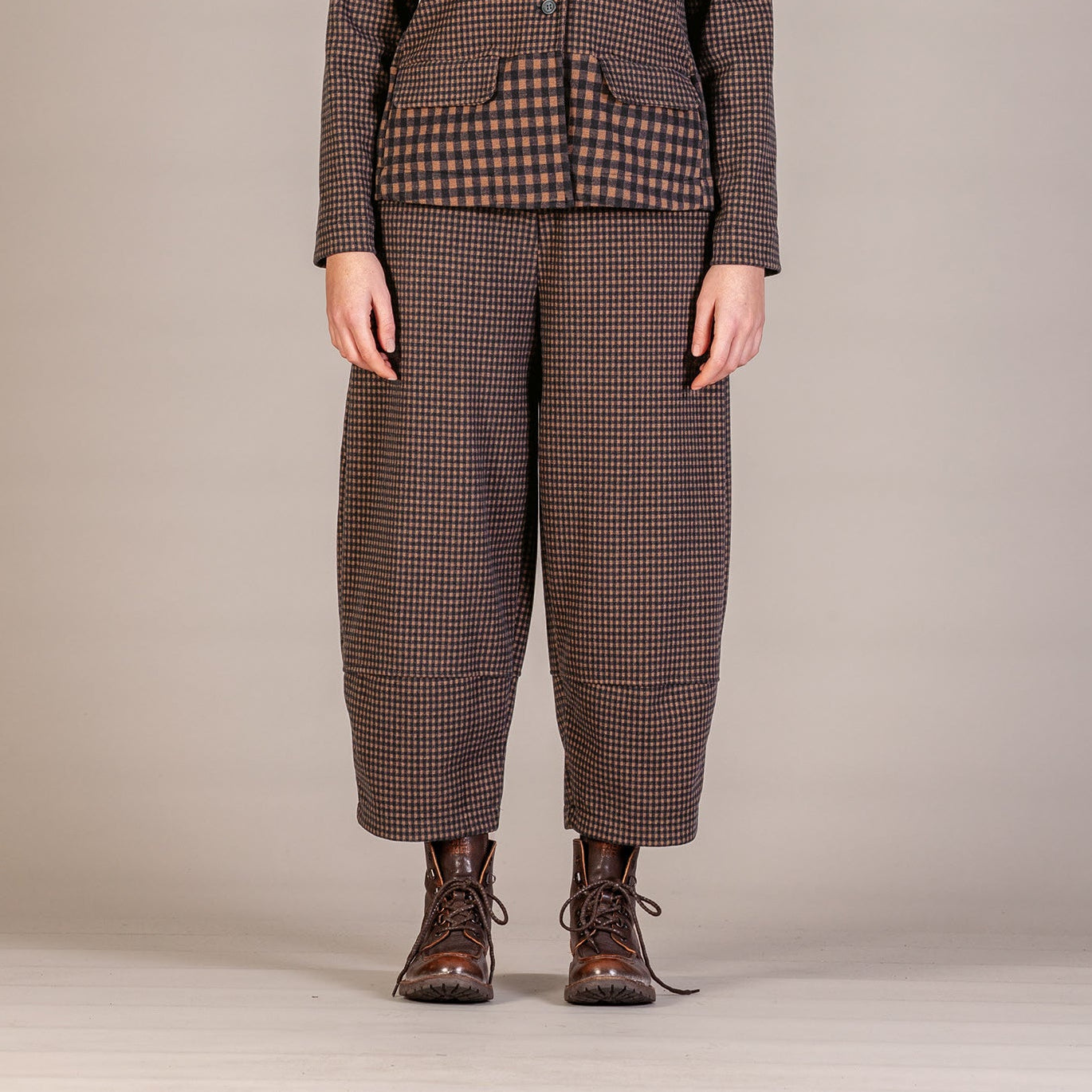 A model stands facing forward in a studio setting, wearing a vintage-style brown and black checkered jacket and matching wide-leg trousers, paired with a dark blue mock-neck shirt and brown lace-up boots.