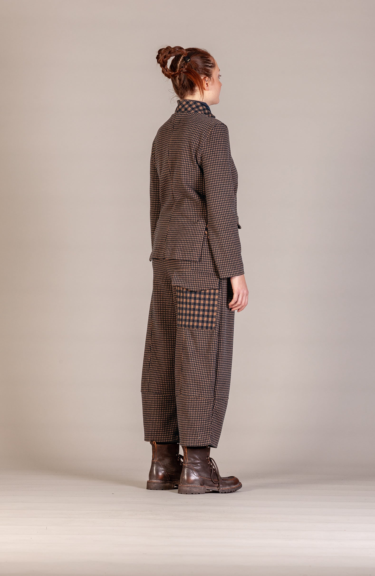 A model shown from the back wearing a brown and black checkered blazer and matching wide-leg trousers with a patterned patch pocket, paired with dark brown leather boots, against a neutral beige studio background.