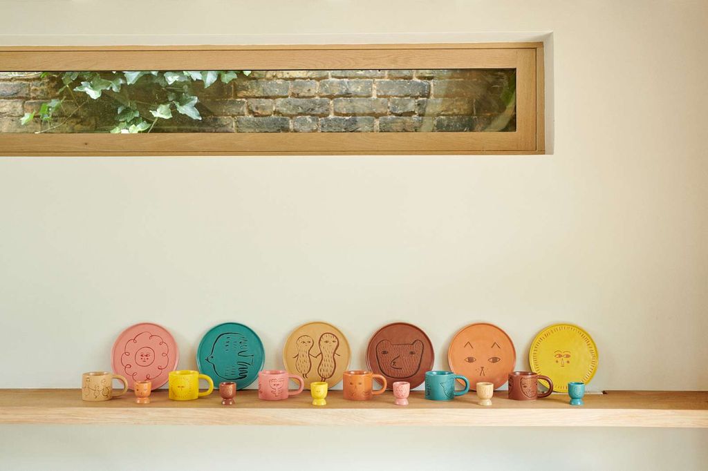 A row of colorful ceramic tableware, including plates and small cups, arranged on a wooden shelf against a cream wall with a window above showing a brick wall and greenery.