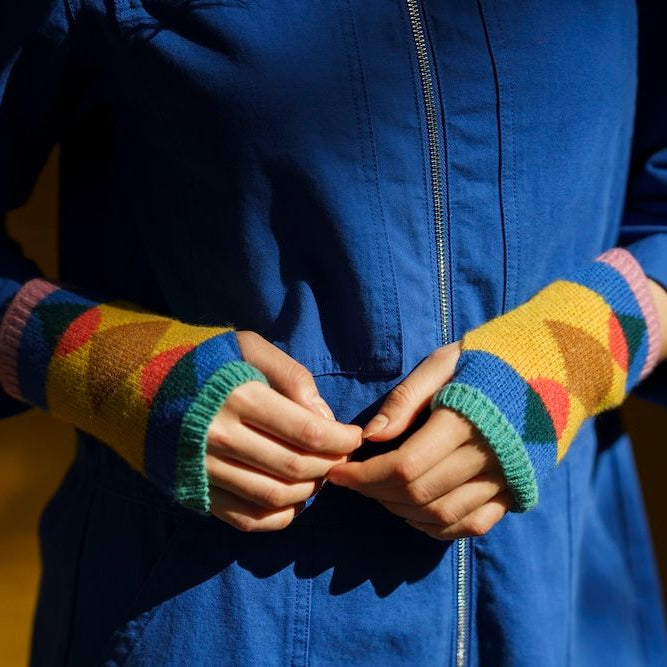 A close-up of a person wearing a bright blue zip-front jacket and colorful knit fingerless wrist warmers with a geometric pattern in yellow, green, pink, orange, and brown.