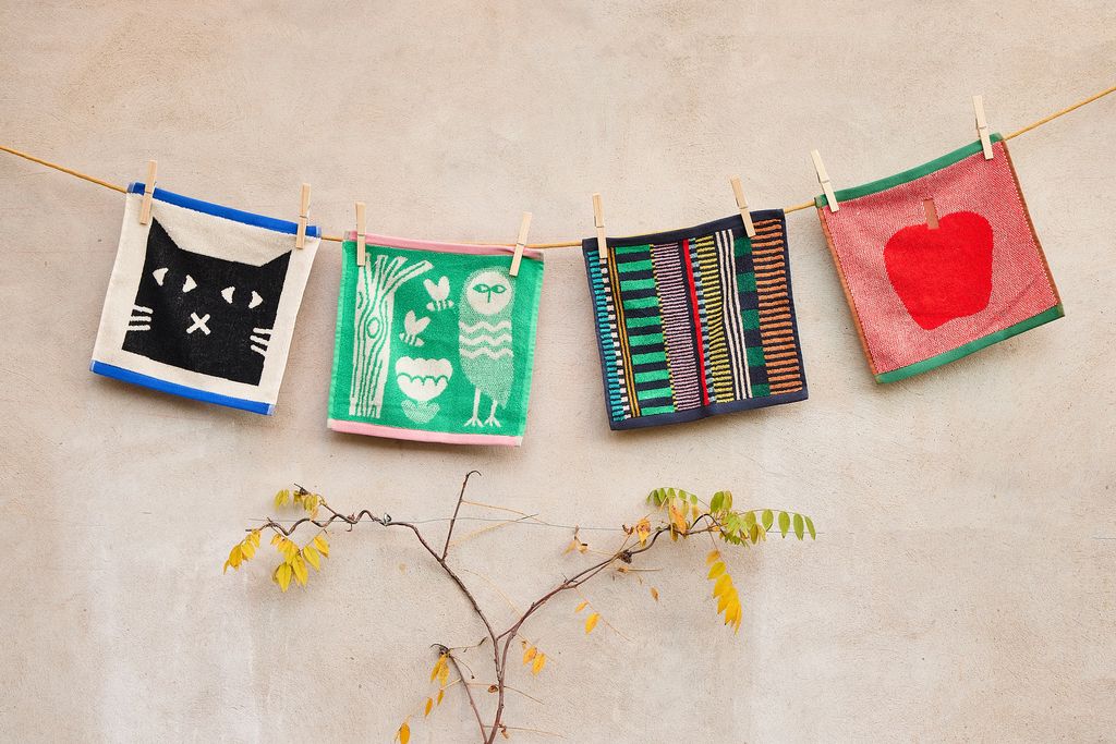 Four colorful woven fabric napkins hanging from a string with wooden clothespins against a textured beige wall, with a small branch and yellow leaves at the bottom.
