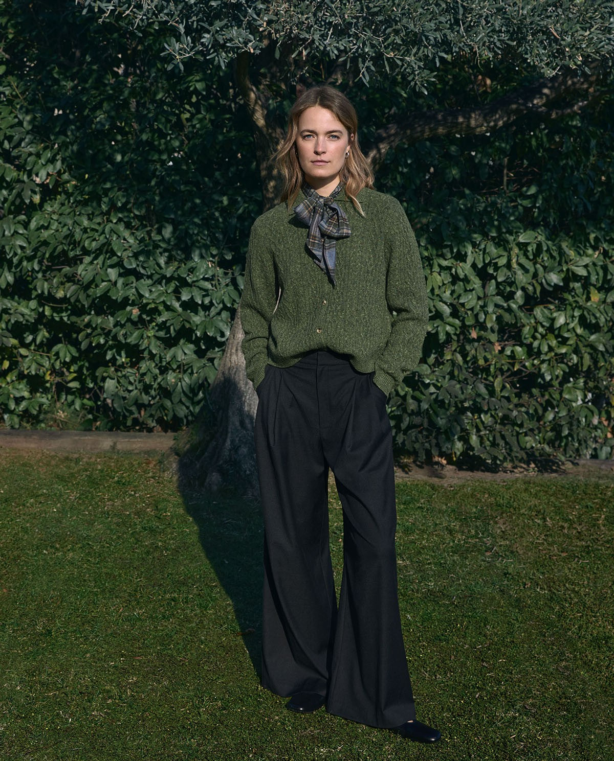 A woman stands outdoors on grass in front of a leafy hedge, wearing an olive green textured knit sweater, black wide-leg trousers, and a dark patterned neck scarf.