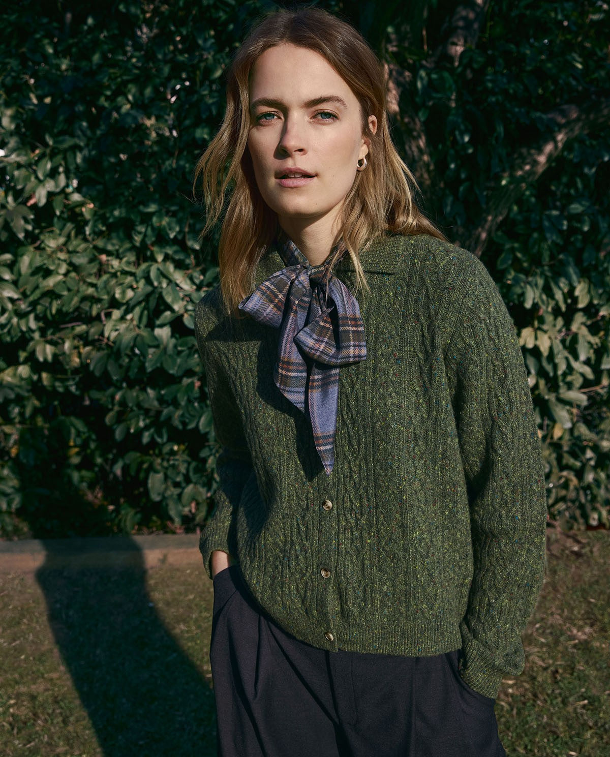 A woman standing outdoors in front of dense green foliage, wearing an olive green cable-knit button-up cardigan with a blue and gray plaid bow tie-neck blouse underneath and dark trousers.