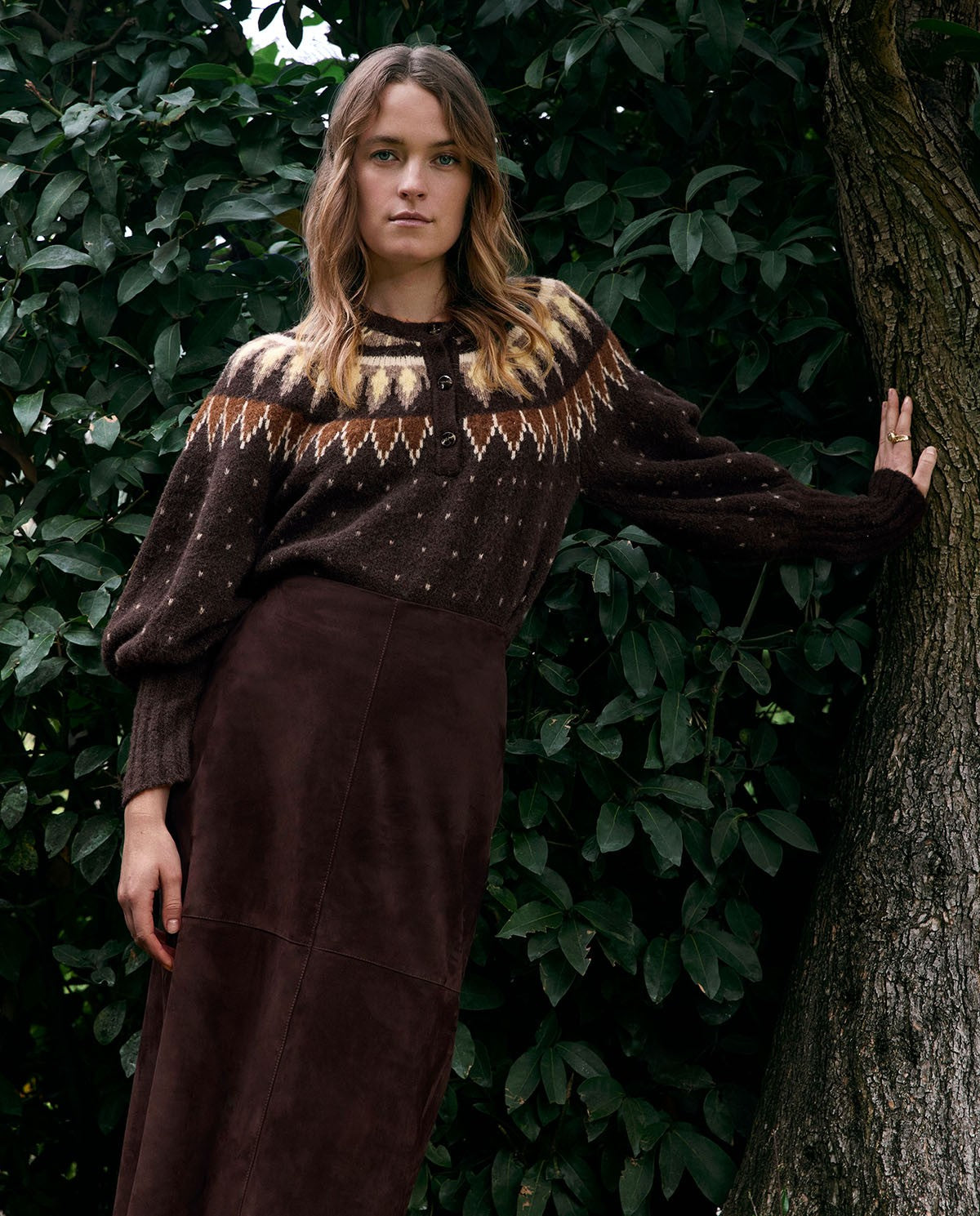 A woman stands outdoors against a backdrop of dark green foliage and a tree trunk, wearing a dark brown knit sweater with a cream and tan Fair Isle yoke pattern and a matching dark brown maxi skirt.