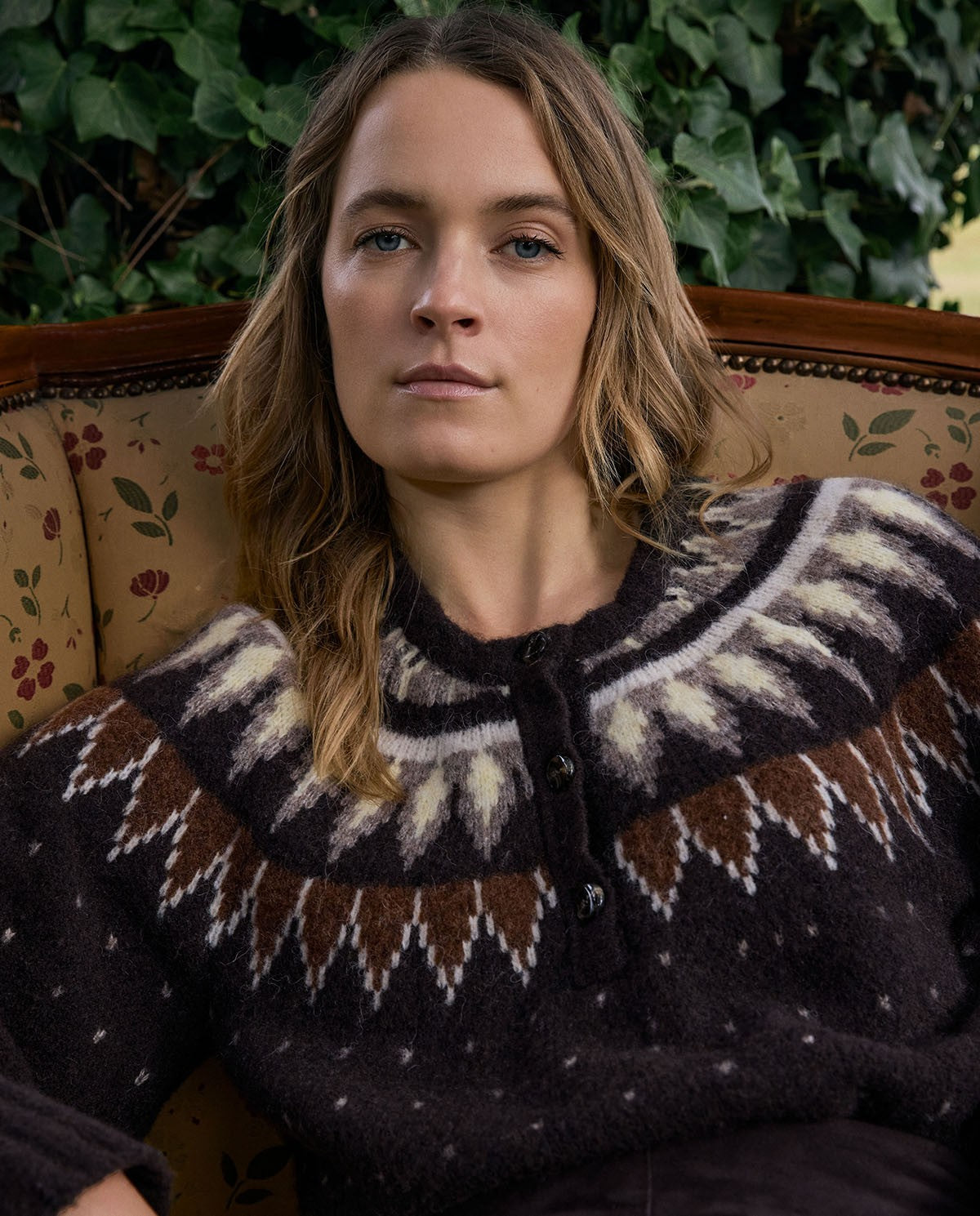 A woman wearing a dark brown knit sweater with a cream and brown geometric pattern around the collar, sitting on a floral armchair with green ivy in the background.