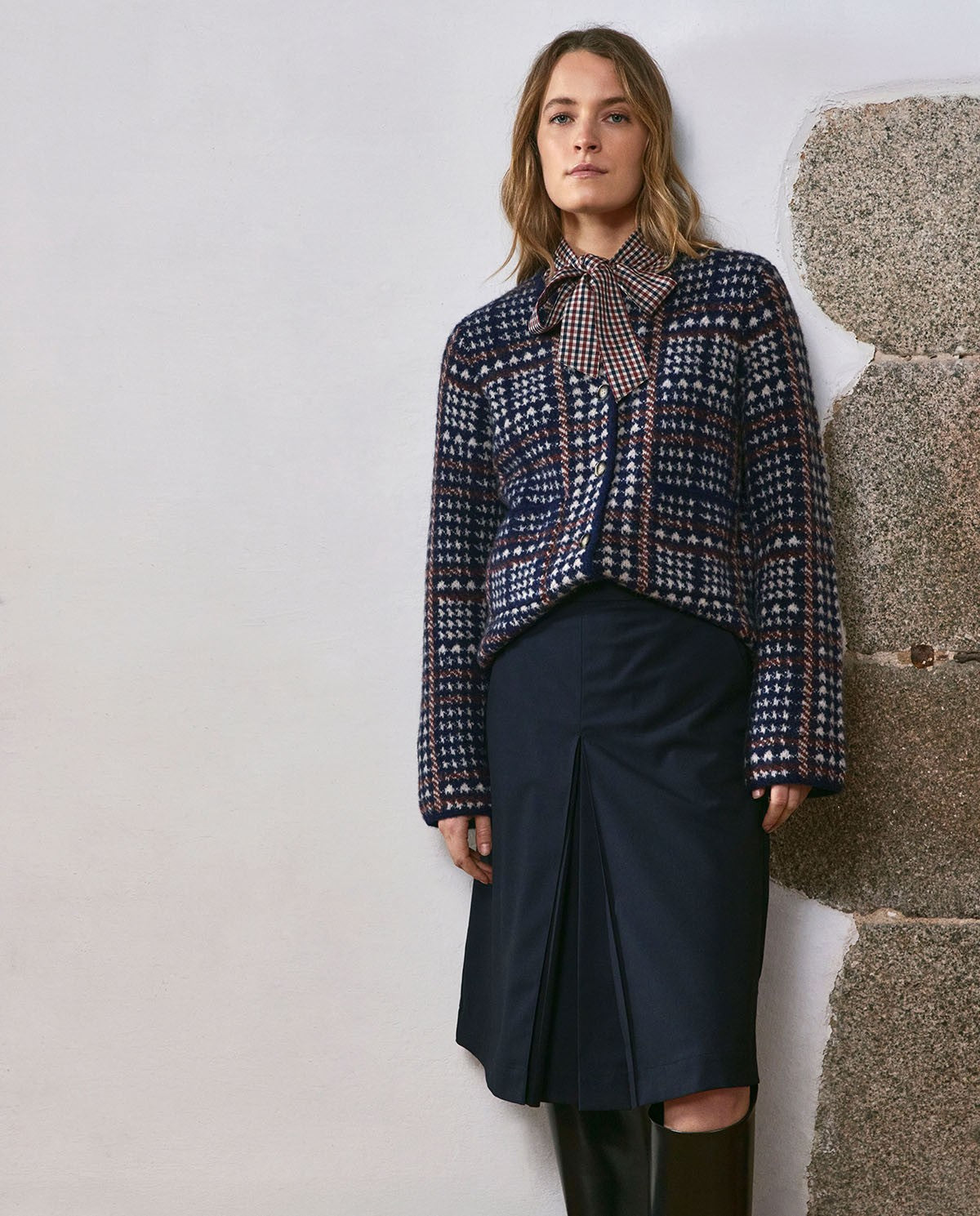 A woman stands against a white wall with rough stone on the right, wearing a patterned navy, red, and white cropped jacket with a bow tie collar over a dark navy skirt.