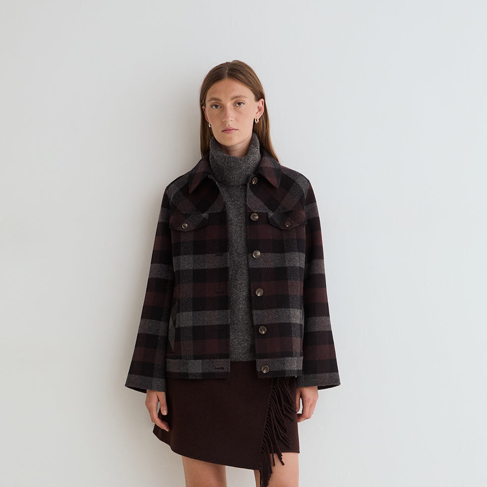 A model stands against a plain white studio background wearing a dark plaid button-front jacket over a gray turtleneck, a dark brown fringe mini skirt, and black knee-high boots.