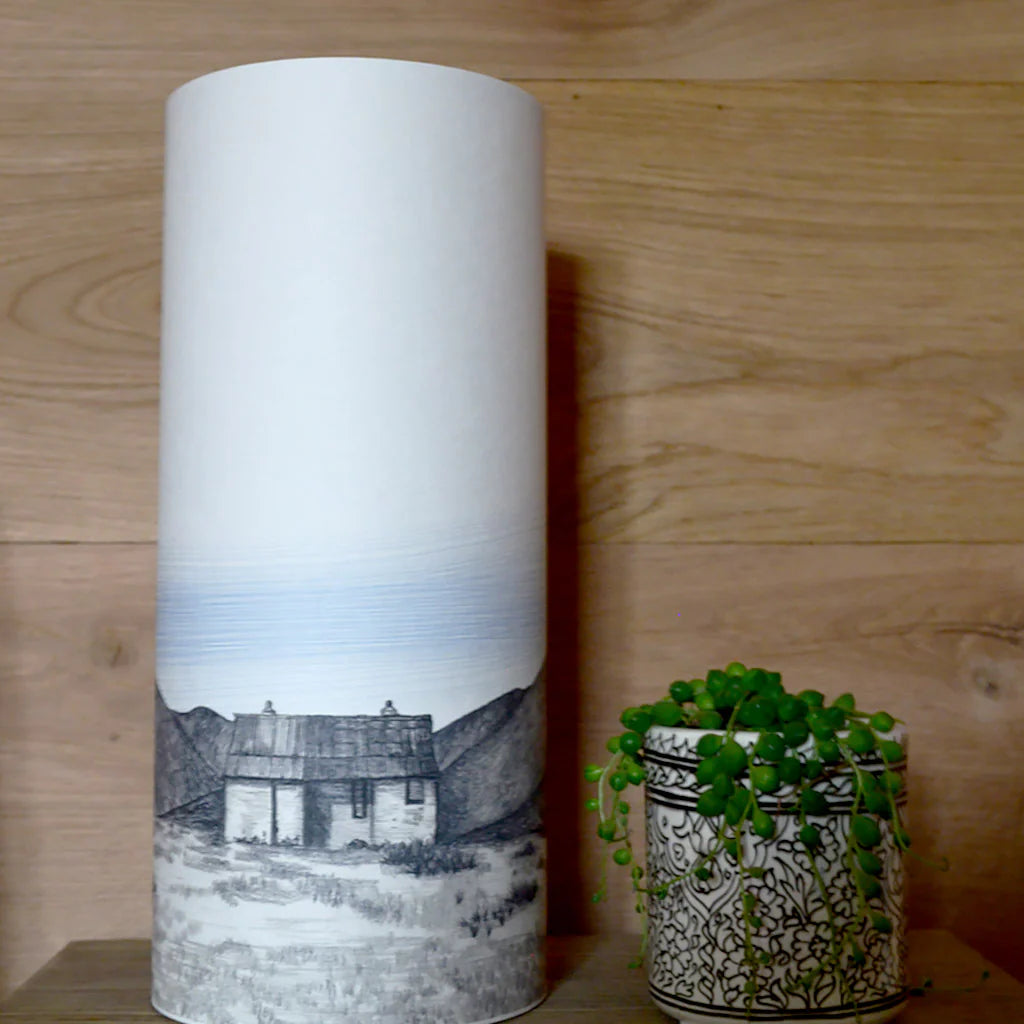 Tall cylindrical lampshade with a grayscale illustration of a cottage in a mountainous landscape.