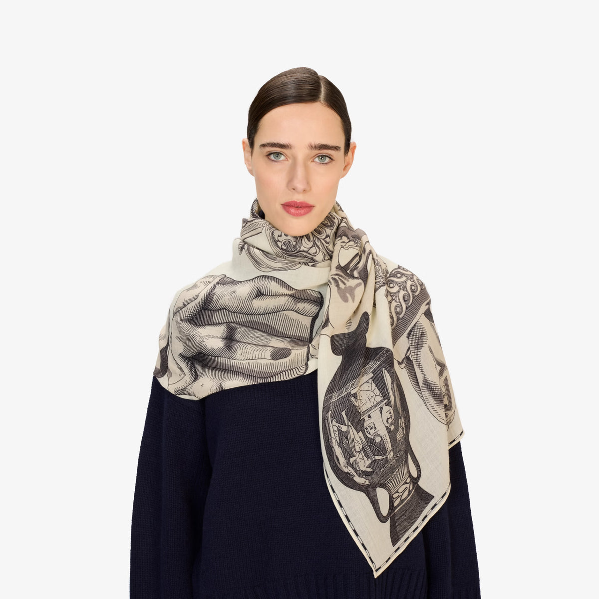 A model wearing a dark navy sweater and a beige scarf with a black graphic print of hands and a classical urn.