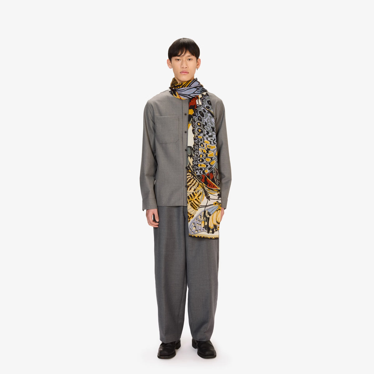 A model stands against a plain white studio background wearing a gray three-piece outfit consisting of a long-sleeve button-up shirt, matching trousers, and a patterned scarf with black, gray, yellow, and orange details.