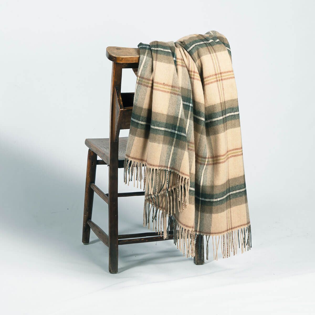 Beige and green plaid throw blanket with fringe, draped over a wooden chair.