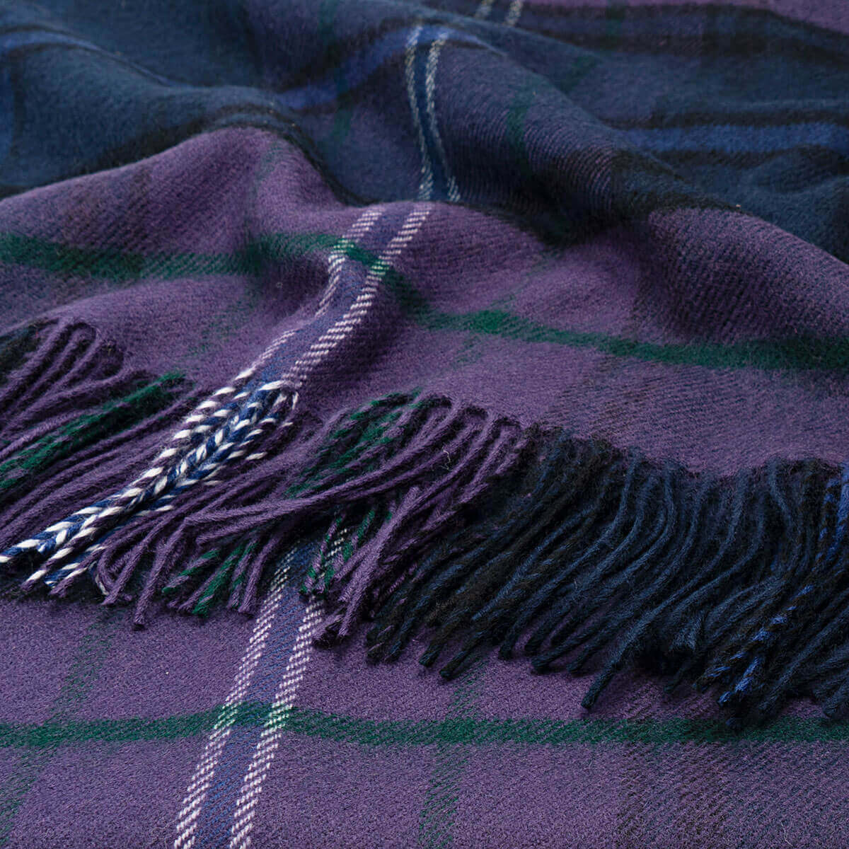 A close-up of a plaid fringed scarf or throw in deep purple with green and blue lines, featuring braided tassels.