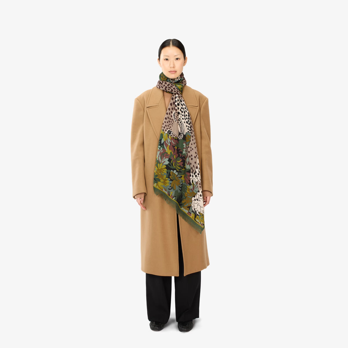 A model stands against a light gray studio background wearing a long camel coat, black wide-leg trousers, and a patterned scarf with leopard print and floral elements.
