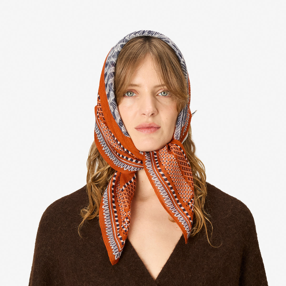 A woman wearing a patterned orange headscarf with black, white, and gray geometric designs, styled with a brown knit sweater against a plain white background.