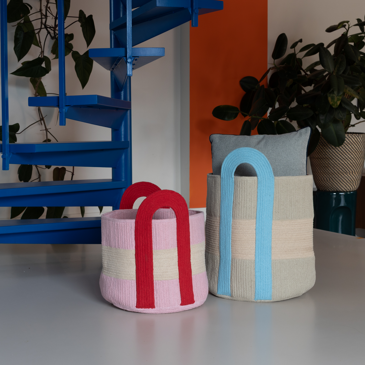 Two colorful woven baskets on a table with a blue shelf and plants in the background.