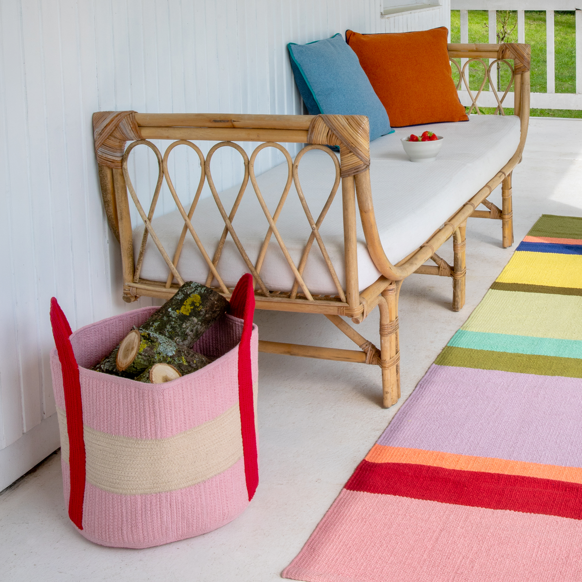 A pink and beige striped woven basket with red handles sits on a white porch floor next to a bamboo daybed with colorful pillows and a striped rug.