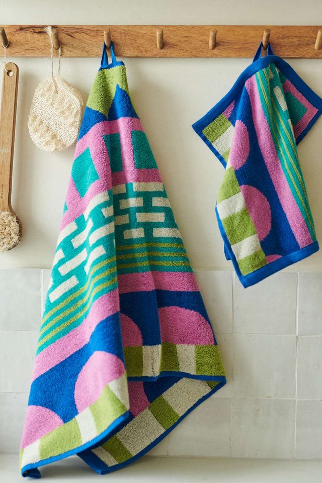 Two colorful patterned towels hanging from a wooden peg rack on a tiled wall. The towels feature bold geometric shapes in blue, pink, green, and white.