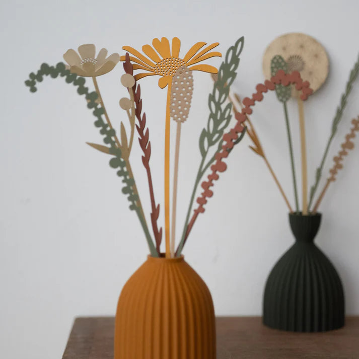 A decorative dried floral arrangement in a ribbed mustard-yellow vase, displayed on a wooden cabinet against a light wall, with a second dark vase blurred in the background.