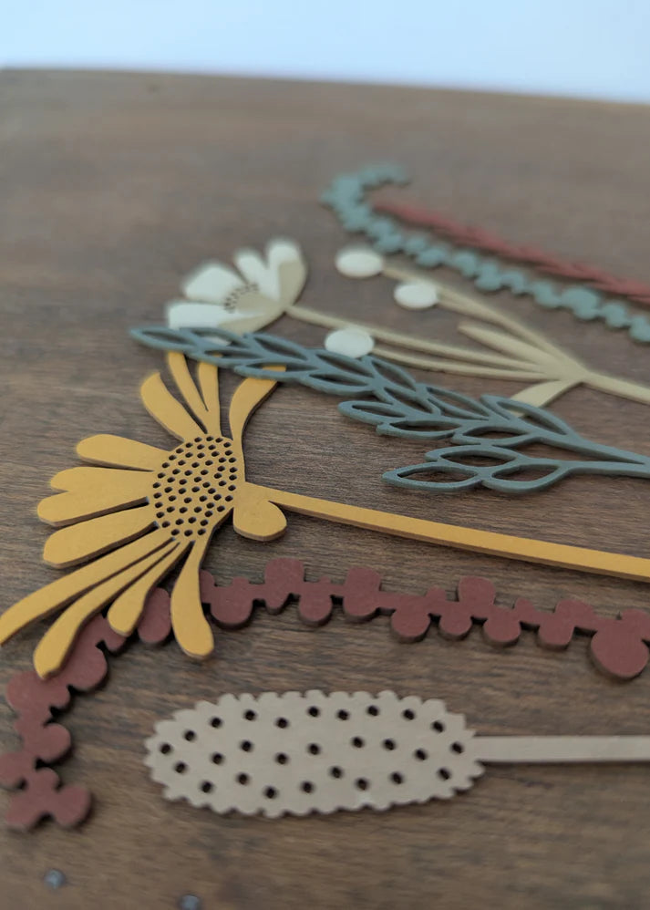 A close-up of layered wooden craft embellishments or scrapbook pieces arranged on a brown wood surface, including a yellow flower, pale green leafy stems, cream blossoms, and dusty rose scalloped floral pieces.