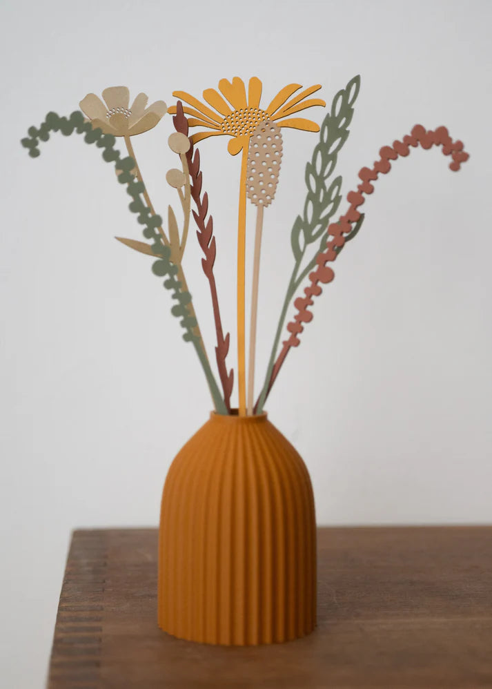 A decorative dried flower arrangement in a ribbed terracotta-colored vase, displayed on a wooden surface against a plain light gray background.