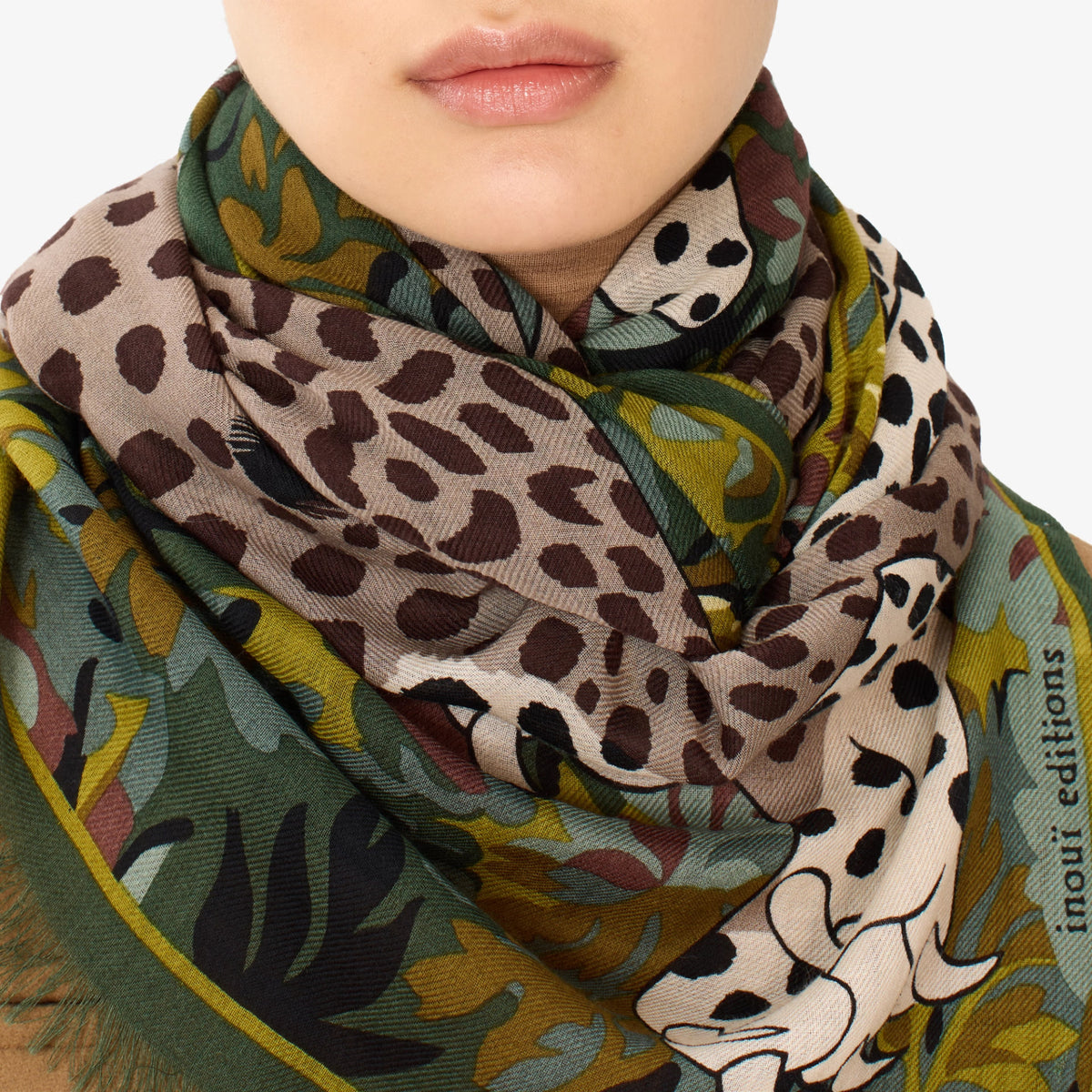A close-up of a model wearing a patterned scarf with a leopard print and botanical design. The scarf features a beige base with dark brown spots, olive green leaves, and yellow accents, with the brand name 'inout editions' printed on the edge.