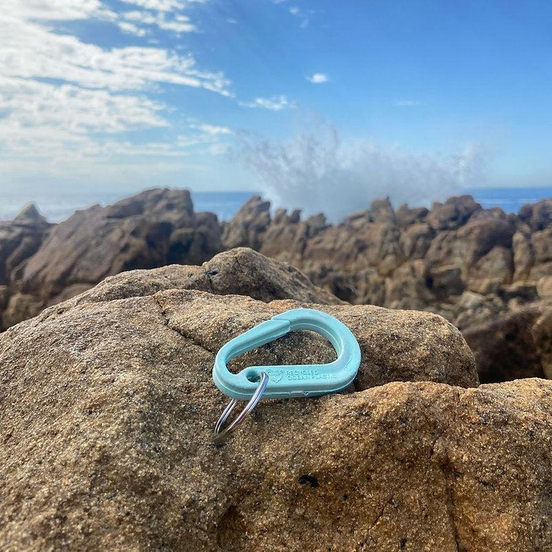 Light blue plastic carabiner keychain rests on a rock.  The carabiner is marked "Recycled Ocean Plastic".