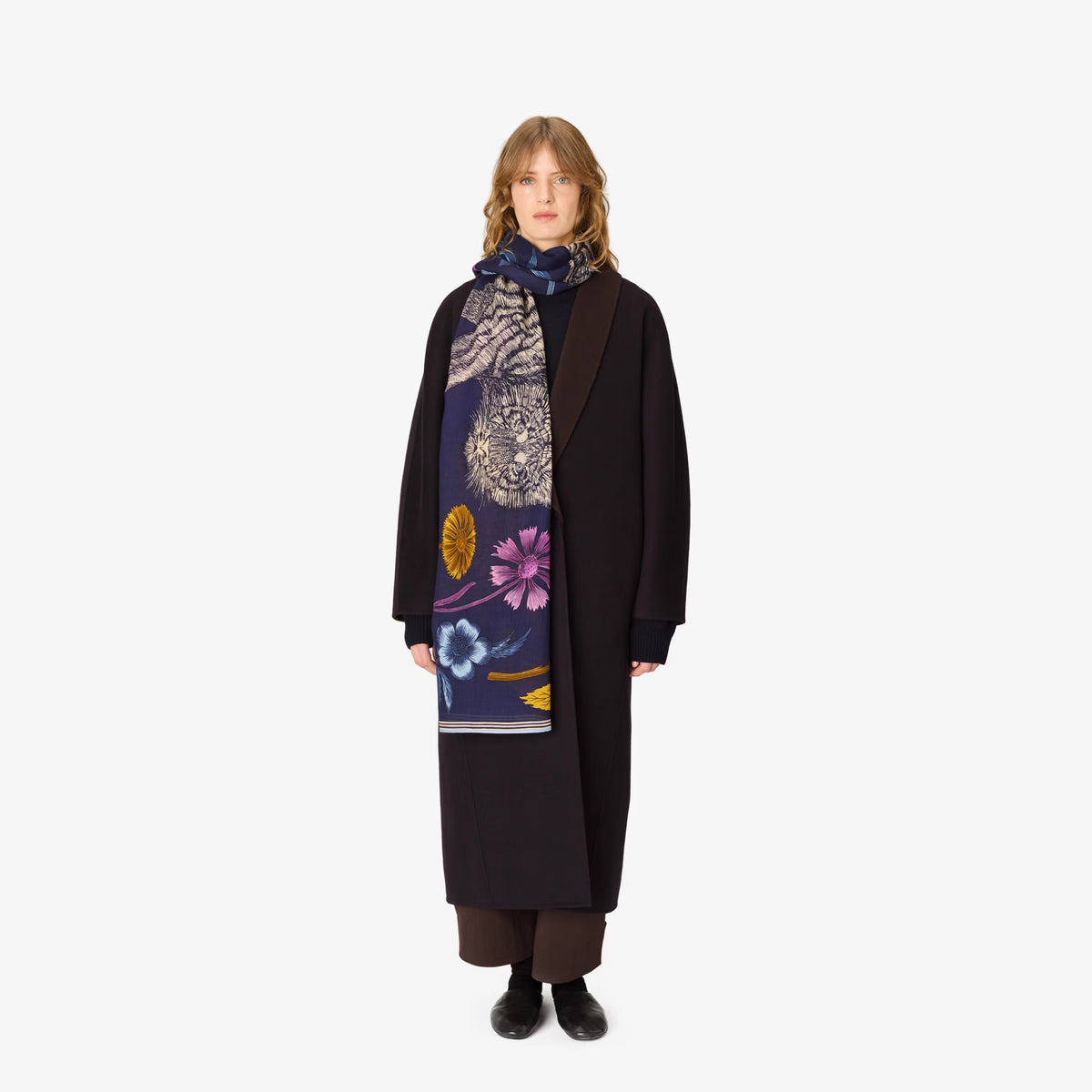 A model stands against a plain white background wearing a long black coat over a navy blue scarf with a colorful floral and abstract pattern, paired with brown trousers and black shoes.