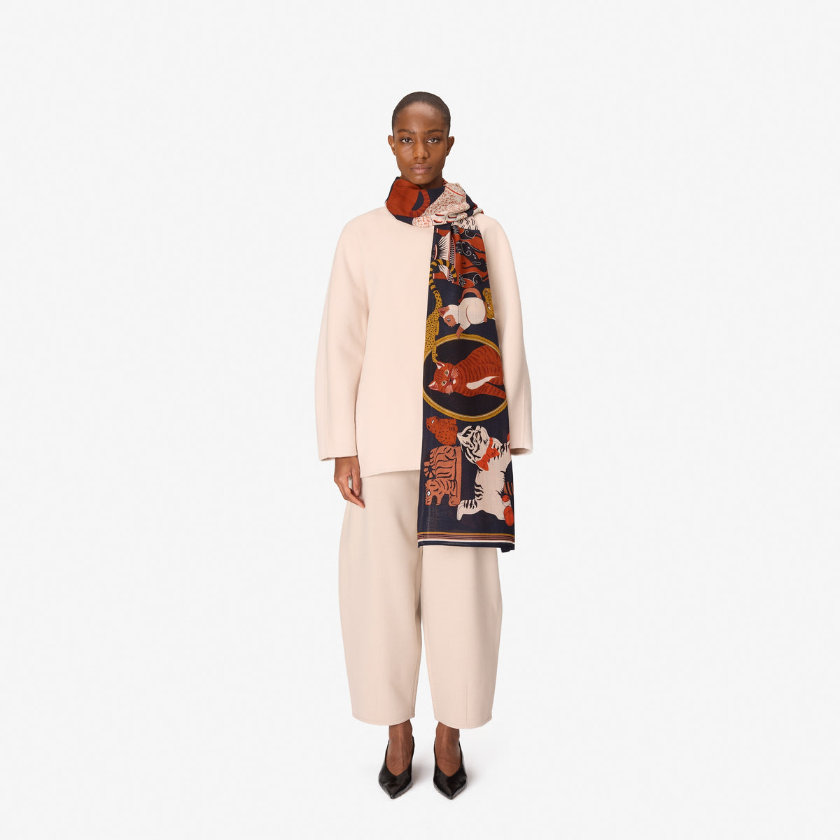 A model stands against a plain white studio background wearing a cream-colored two-piece outfit with a patterned scarf. The scarf features illustrations of cats in orange, red, black, and white, wrapped around the neck over a rust-colored inner layer.