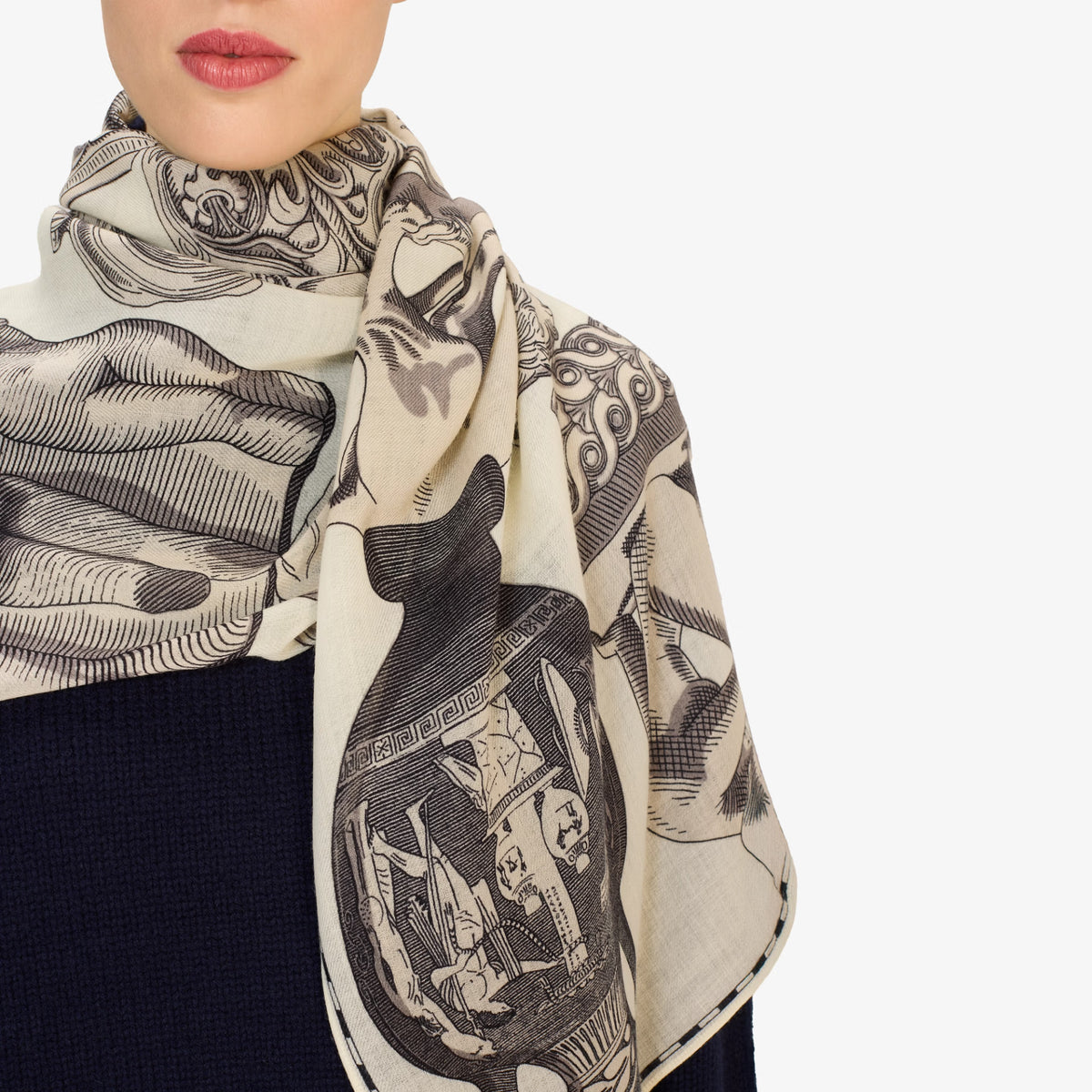 A close-up of a model wearing a square scarf with a black and beige graphic print featuring a cat and abstract patterns, paired with a dark navy top against a light gray background.