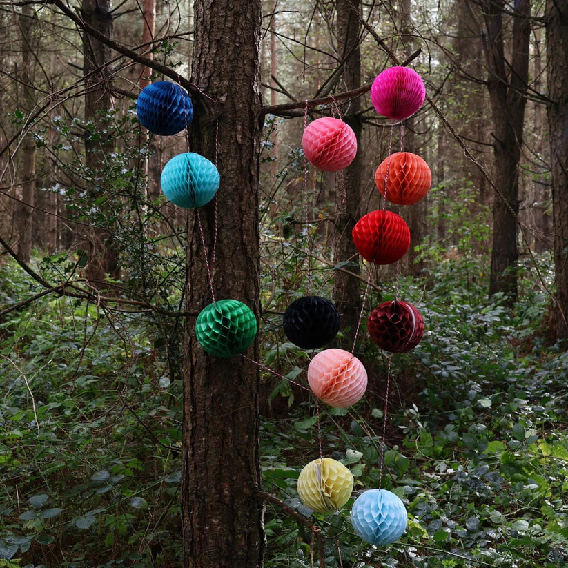 String of colorful honeycomb paper balls hanging from a tree in a forest.