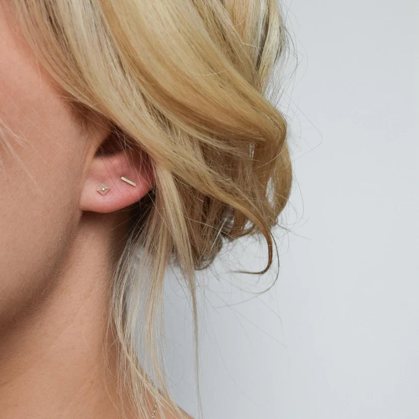 Delicate gold stud earrings; one small geometric shape, the other a thin bar.