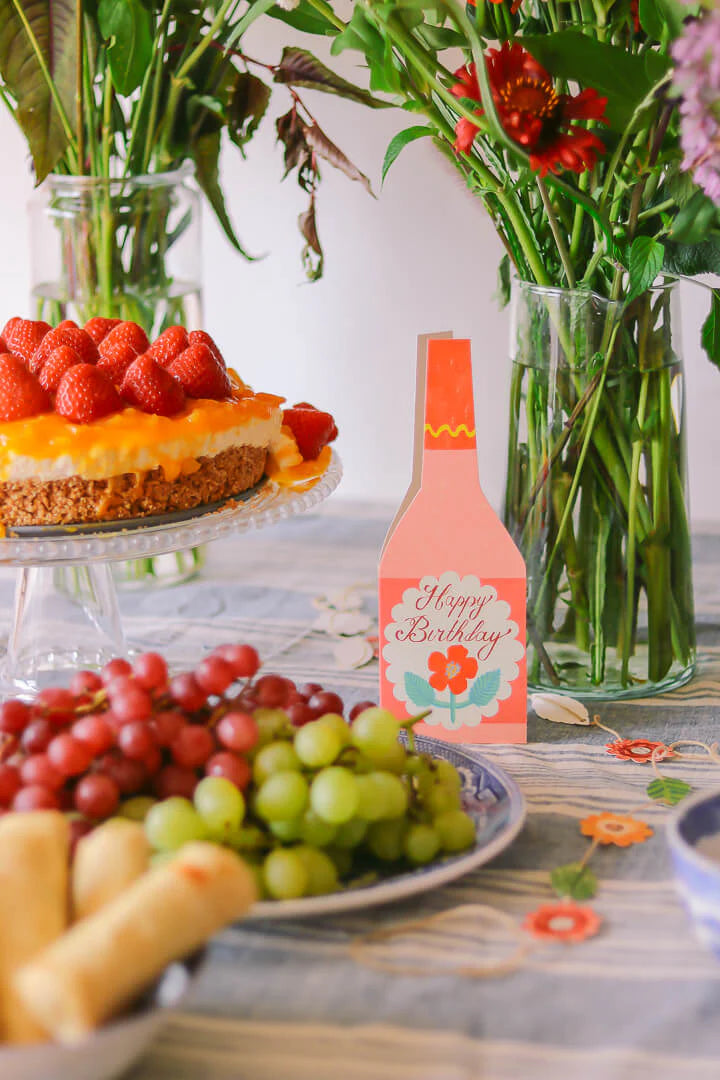 Strawberry mango cheesecake on a cake stand, next to a "Happy Birthday" card and bowls of grapes.