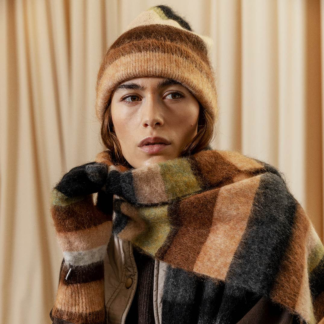 Person wearing a striped beanie and scarf against a beige curtain background
