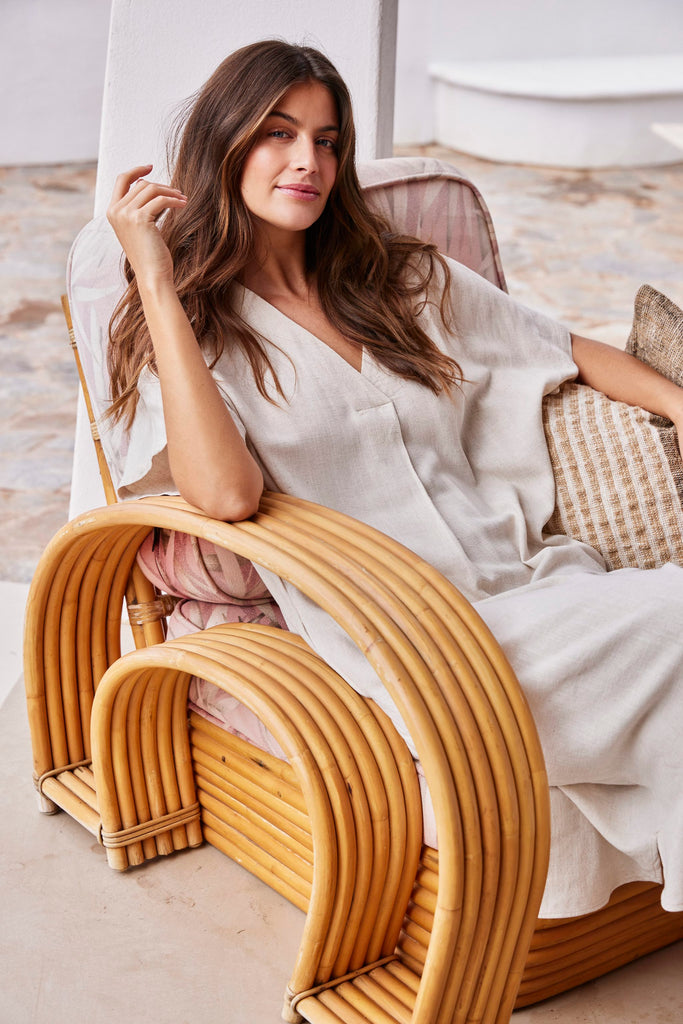 A woman in a light beige Fransa outfit sits on a rattan chair, smiling, with soft, natural tones around her.