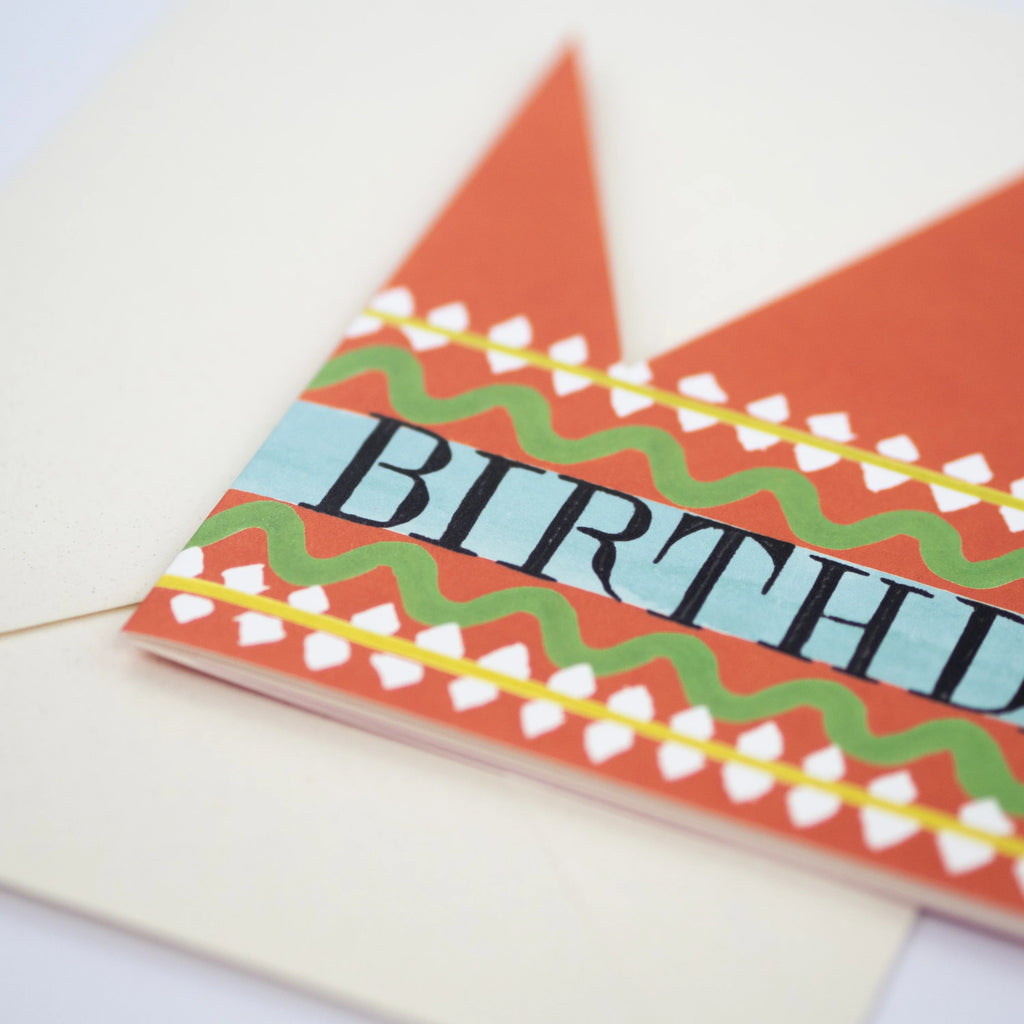 Orange birthday card shaped like a crown; features a teal band with "BIRTHDAY" in black lettering and decorative green and