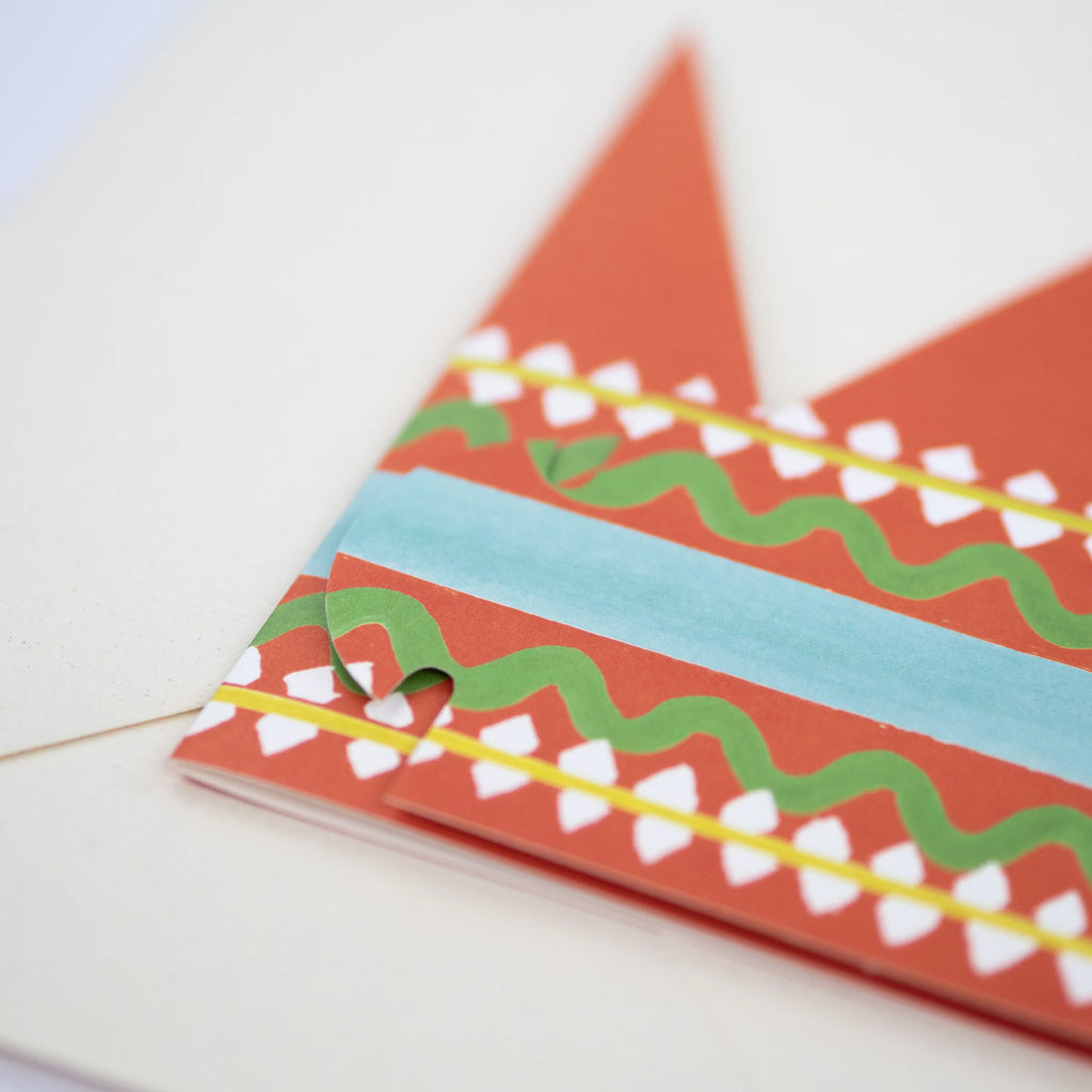 A pop-up card partially unfolded, featuring a red, green, and teal patterned design.  The card is shaped like a crown.