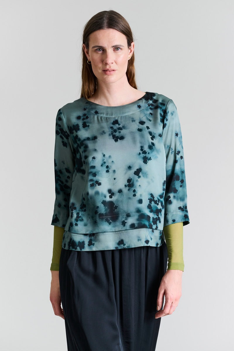Woman wearing a green and blue patterned top with black pants on a white background