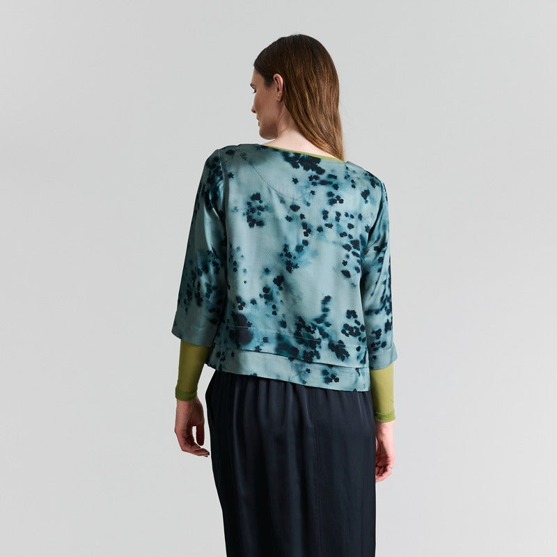 Woman wearing a green and blue patterned top with black pants on a gray background