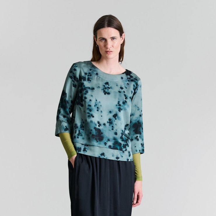 Woman wearing a teal and black patterned top with green sleeves on a light gray background
