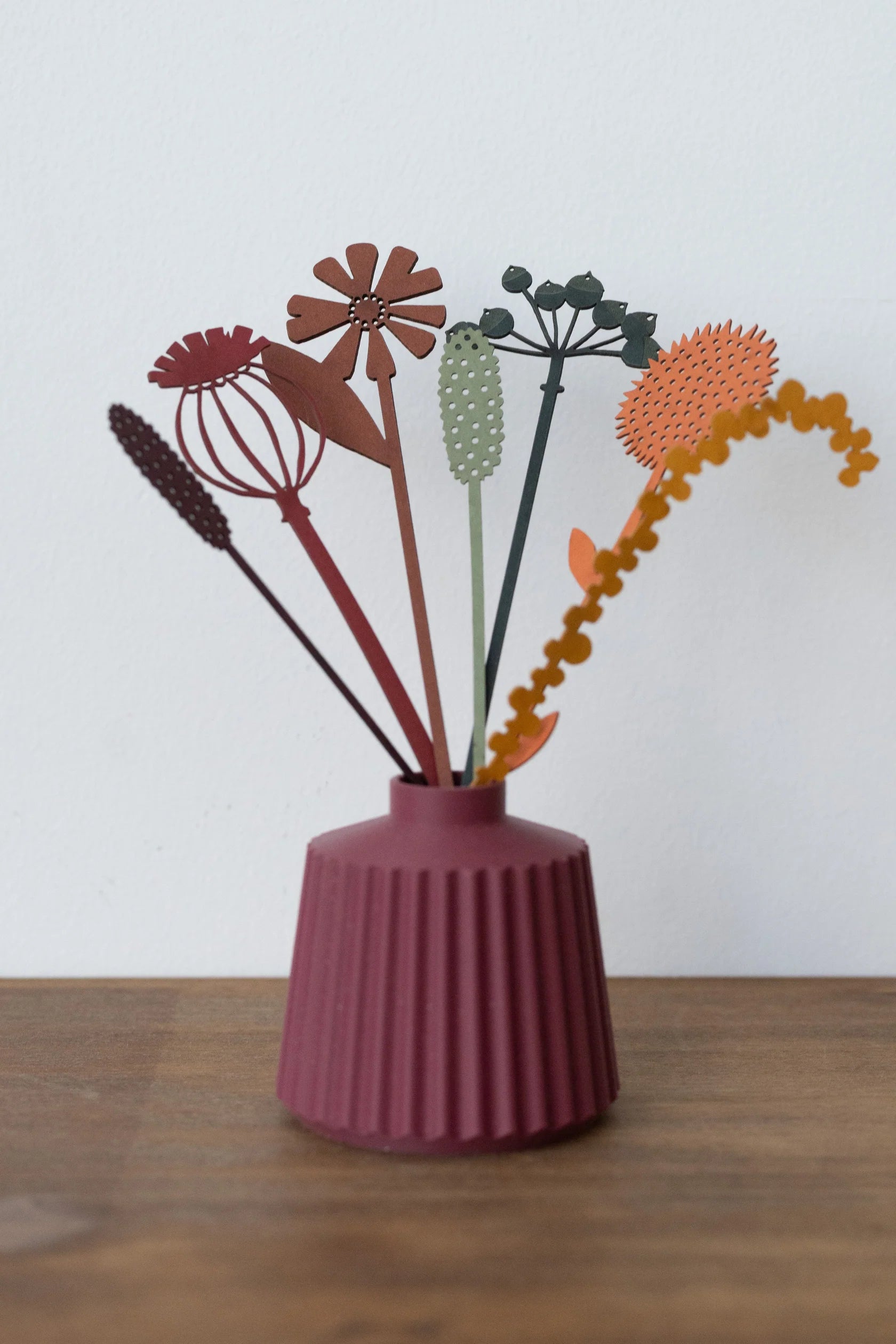Reddish-brown vase with fluted sides holding a bouquet of stylized, multicolored paper flowers and stems.
