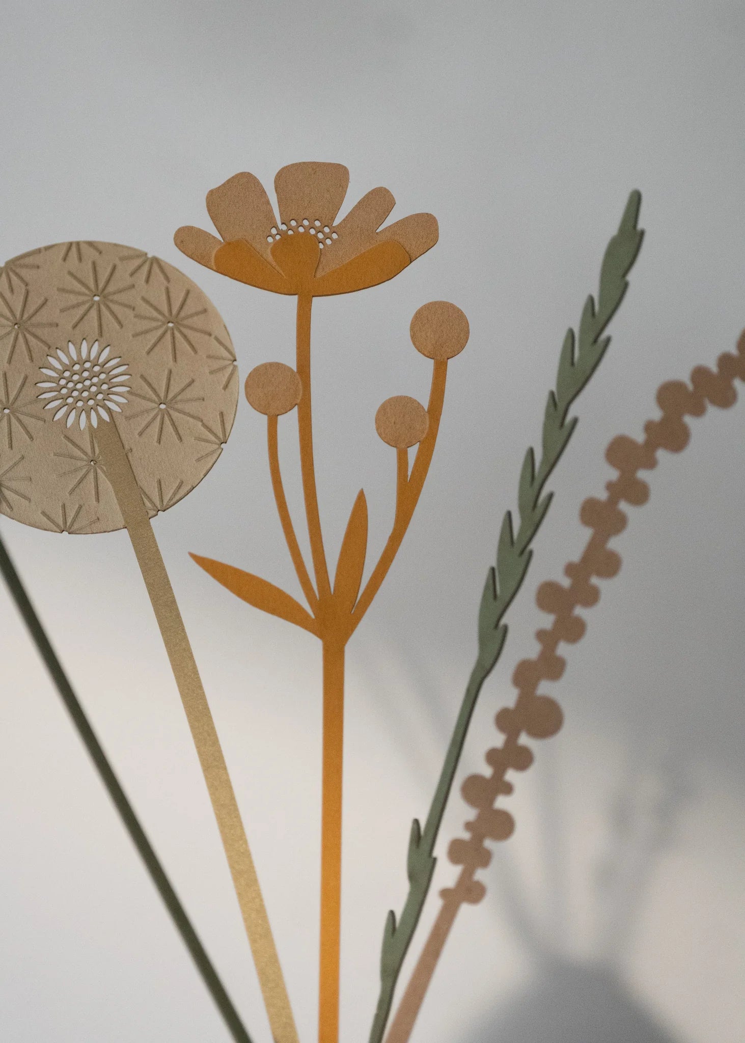 Paper cut-out floral arrangement featuring a dandelion, flower, and grasses in muted orange and green tones.