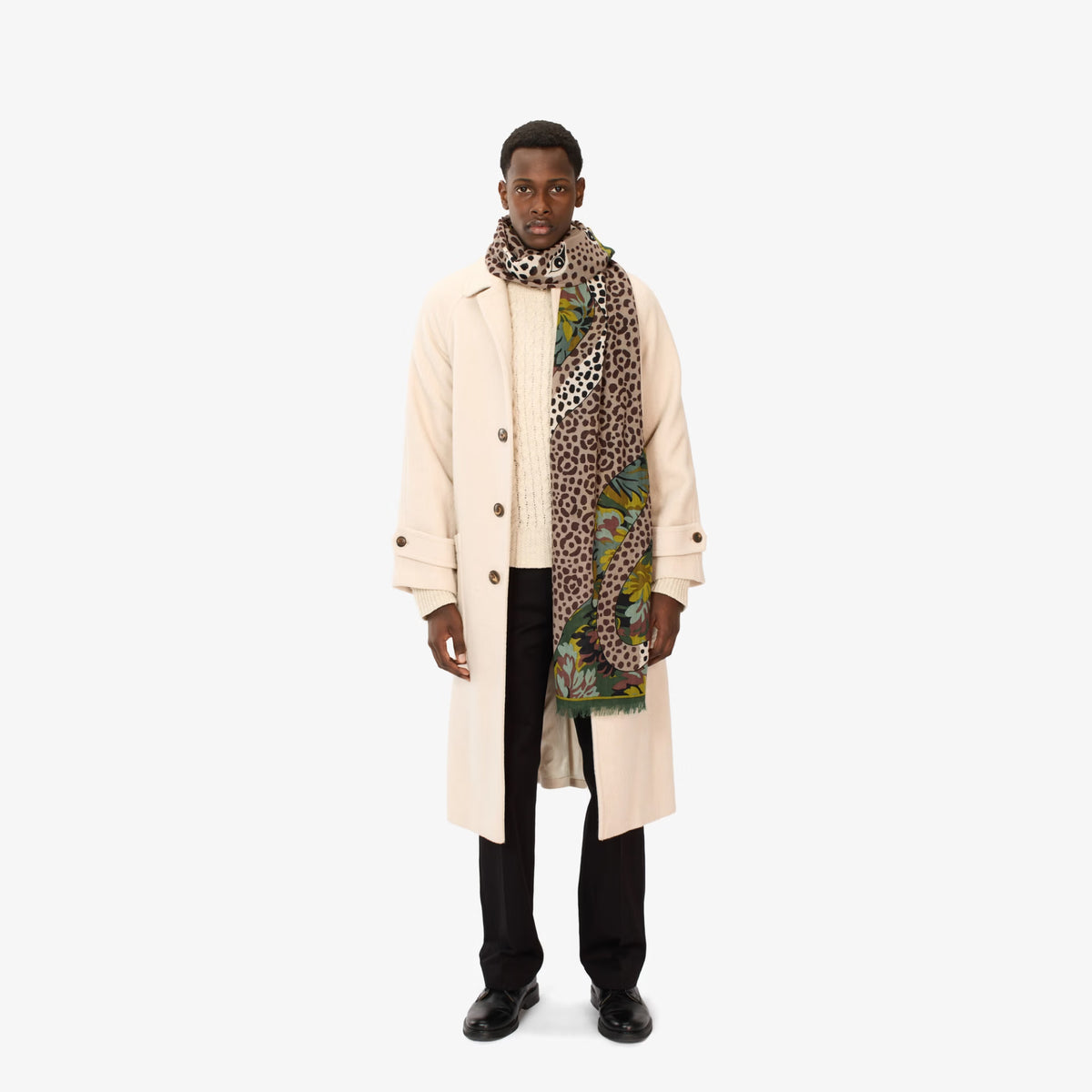 A male model stands against a plain light gray studio background wearing a long cream coat over a cream knit sweater, black trousers, and black shoes, with a patterned scarf wrapped around his neck. The scarf has a leopard print base with green, yellow, and orange abstract graphic elements.