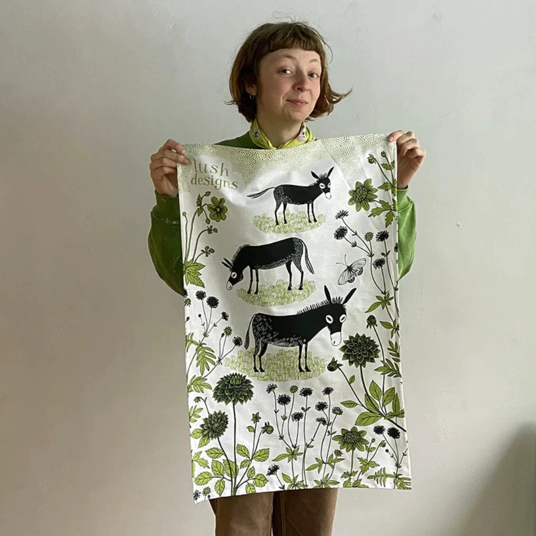 Lush Designs tea towel featuring a repeating pattern of black donkeys and green foliage on a white background.