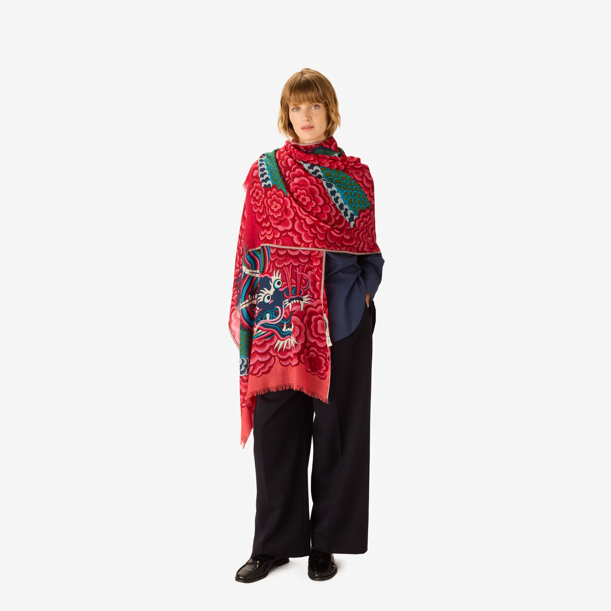 A model stands against a light gray studio background wearing a vibrant red shawl with intricate green, blue, and white floral and dragon motifs, paired with dark navy wide-leg trousers and black shoes.