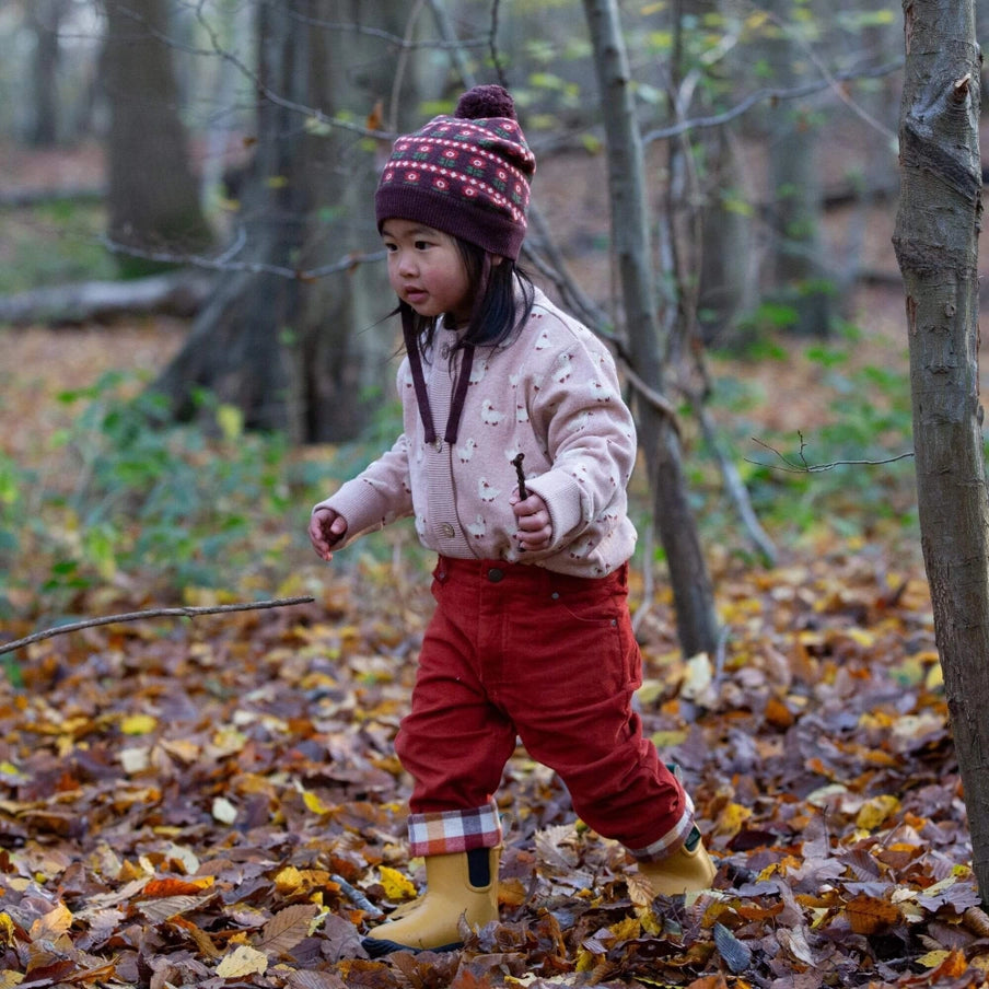 Child in a forest wearing a pink jacket, red pants, and yellow boots, holding a stick.