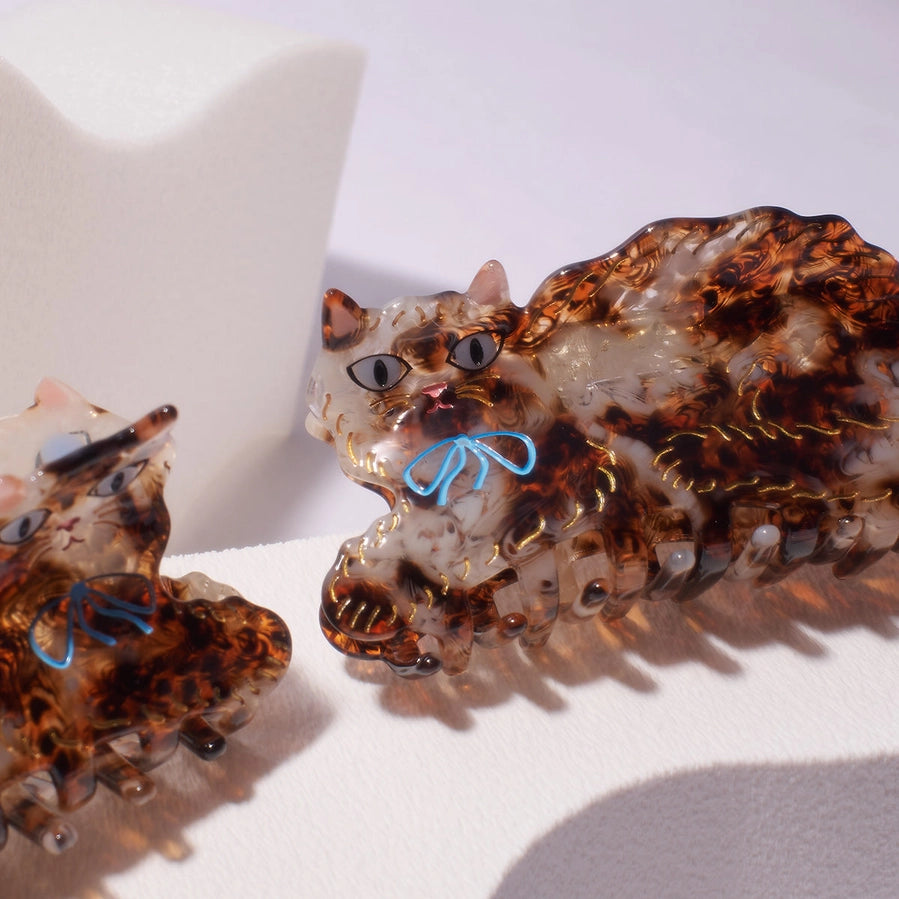 A close-up of a decorative cat-shaped hair claw clip made from translucent amber-brown and white resin with gold glitter details and a small blue bow, shown on a white surface.